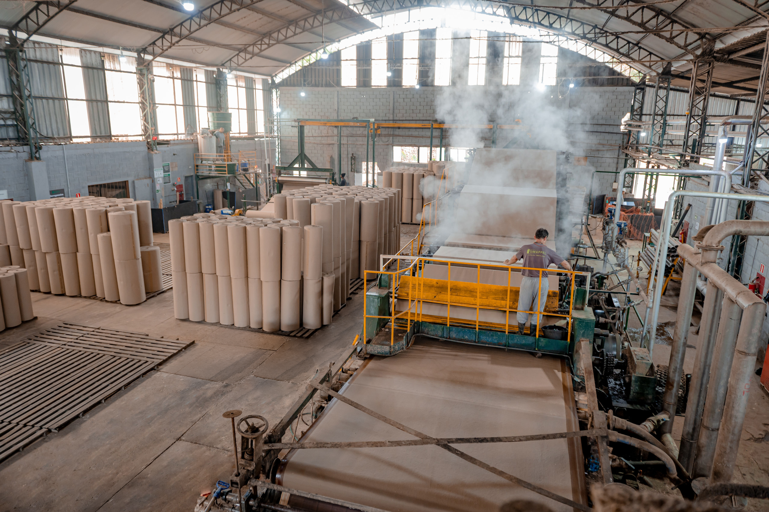 Vista aérea do interior de uma fábrica de papel, mostrando uma grande máquina de produção de chapas onduladas em operação